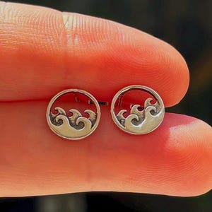 May include: A pair of small, circular silver stud earrings. Each earring features a cutout design depicting stylized ocean waves within the round frame. The metal has a polished finish with darkened recessed areas to highlight the wave pattern.