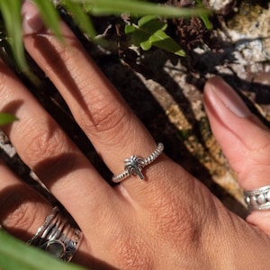 May include: A silver ring with a palm tree design. The ring is on a person's finger, and there are other rings on the other fingers.