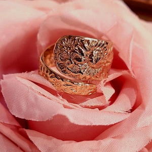 May include: A rose gold-toned metal ring featuring an adjustable wrap-around design. The surface of the ring is embossed with a detailed floral pattern, including visible petals and leaves, creating a textured finish across the entire band.