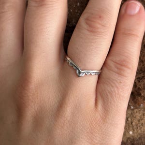 May include: A silver-colored ring with a V-shaped design. The ring has a delicate, patterned edge along the top and bottom of the V. The ring is on a hand with a brick background.