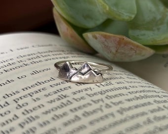 Könnte beinhalten: Ein silberner Ring mit einem Bergketten-Design. Der Ring liegt auf einem Buch mit Text.