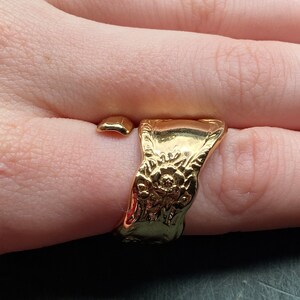 May include: A gold-toned open ring featuring a wide, flat band with an embossed floral pattern. The ring has a polished, reflective surface and a gap between the two ends, creating a wrap-around style that sits on the finger.