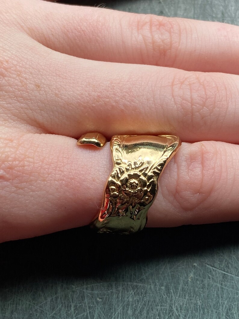 May include: A gold-toned open ring featuring a wide, flat band with an embossed floral pattern. The ring has a polished, reflective surface and a gap between the two ends, creating a wrap-around style that sits on the finger.