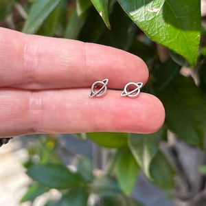 May include: A pair of small, silver-colored planet-shaped stud earrings. Each earring features a circular planet with a thin, curved ring around it. The earrings are displayed against a background of green foliage.