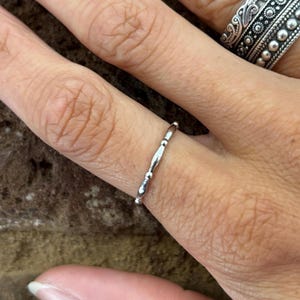 May include: A silver ring with a delicate design featuring alternating smooth and textured sections. The ring is worn on a finger, showcasing its slender profile and polished finish. Another ornate ring is visible in the background.