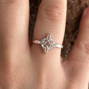 May include: A silver ring with an ornate, symmetrical design. The ring features a central, four-leaf clover-like pattern with delicate, swirling details. The band is thin and polished, and the ring is shown on a finger.