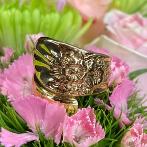 May include: A gold-toned ring with a floral design. The ring is set against a background of pink and green flowers.