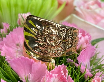 May include: A gold-toned ring with a floral design. The ring is set against a background of pink and green flowers.