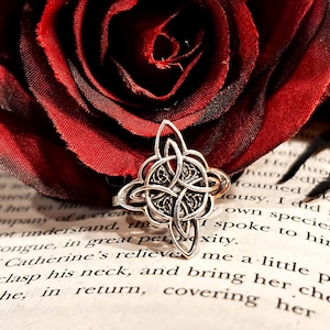 May include: A silver-colored Celtic knot ring with intricate details, set against a backdrop of a deep red rose and a vintage book. The ring's design features interwoven patterns. The background includes text from a book.