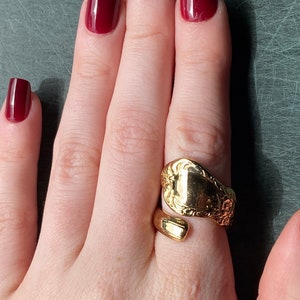 May include: A gold-toned ring crafted from a repurposed spoon handle, featuring an open-ended bypass design. The surface of the ring displays intricate embossed floral detailing on one side, while the other end remains smooth and plain.