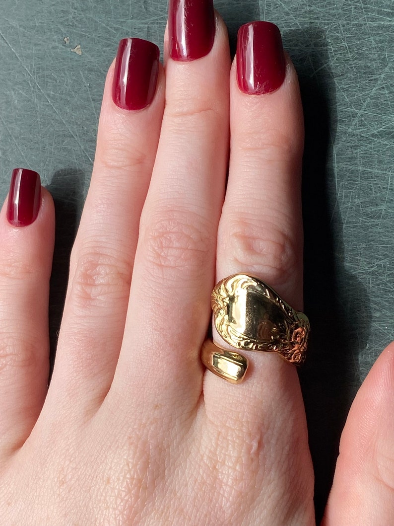 May include: A gold-toned ring crafted from a repurposed spoon handle, featuring an open-ended bypass design. The surface of the ring displays intricate embossed floral detailing on one side, while the other end remains smooth and plain.