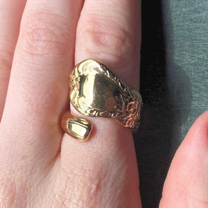 May include: A gold-toned spoon ring featuring an open-ended design. The wide, flat top section displays an intricate, embossed floral pattern along the edges, while the band tapers into a smooth, polished finish that wraps around the finger.