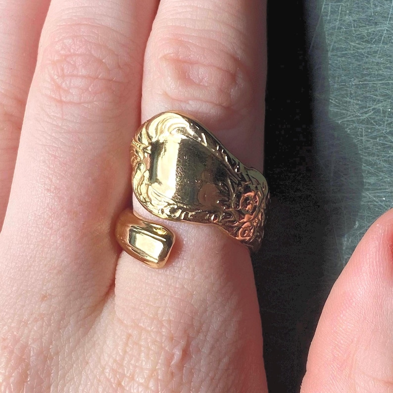 May include: A gold-toned spoon ring featuring an open-ended design. The wide, flat top section displays an intricate, embossed floral pattern along the edges, while the band tapers into a smooth, polished finish that wraps around the finger.