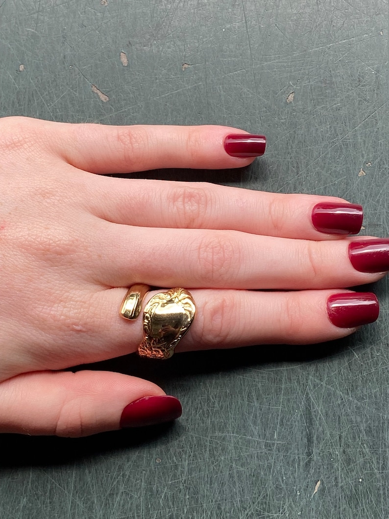 May include: A gold-toned spoon ring featuring an open-ended design. The band is crafted from the handle of a spoon, showcasing an embossed floral pattern on the wider end and a smooth, rounded finish on the narrower, tapering end.