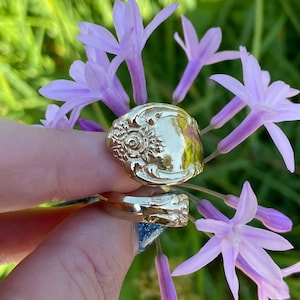 May include: A gold spoon ring with a floral design. The ring is worn on a finger and is surrounded by purple flowers.