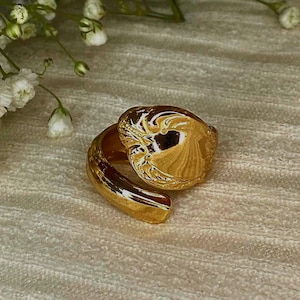 May include: A gold-toned ring featuring a wide, polished band that wraps around to form a rounded, organic shape with a smooth, reflective surface. The ring is positioned near small white flowers with green stems.
