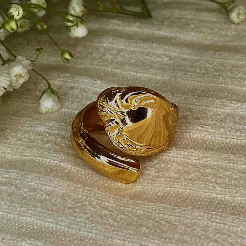 May include: A gold-toned ring featuring a wide, polished band that wraps around to form a rounded, organic shape with a smooth, reflective surface. The ring is positioned near small white flowers with green stems.