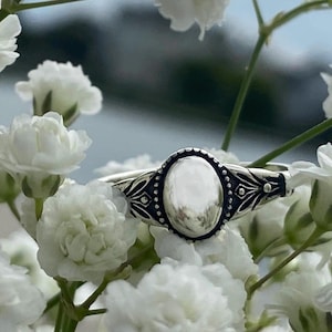 May include: A silver ring with a large oval-shaped stone set in a detailed silver band. The ring is surrounded by white flowers.