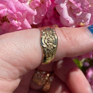 May include: A gold-toned ring with a floral design. The ring is worn on a finger and is surrounded by pink flowers.
