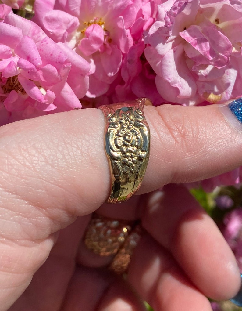 May include: A gold-toned ring with a floral design. The ring is worn on a finger and is surrounded by pink flowers.