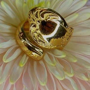 May include: A gold-toned bypass ring featuring a wide, polished band that wraps around the finger. The top section of the ring displays an intricate, embossed pattern with raised details and a reflective, high-shine metallic finish.