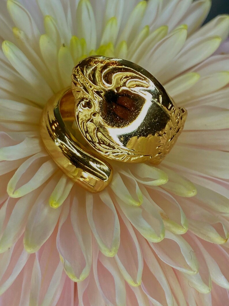 May include: A gold-toned bypass ring featuring a wide, polished band that wraps around the finger. The top section of the ring displays an intricate, embossed pattern with raised details and a reflective, high-shine metallic finish.