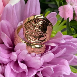 May include: A gold-toned ring with a textured, floral design. The ring is shown on a bed of pink flowers.