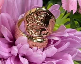 May include: A gold-toned ring with a textured, floral design. The ring is shown on a bed of pink flowers.