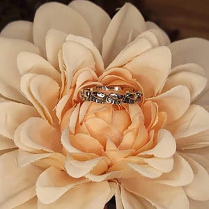May include: A silver ring with an elephant design is displayed on a peach-colored flower. The ring features a repeating pattern of elephant silhouettes. The flower has multiple layers of soft petals, creating a delicate backdrop for the jewelry.