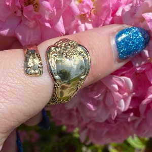 May include: A gold-toned spoon ring with an ornate design. The ring is worn on a finger with blue nail polish.