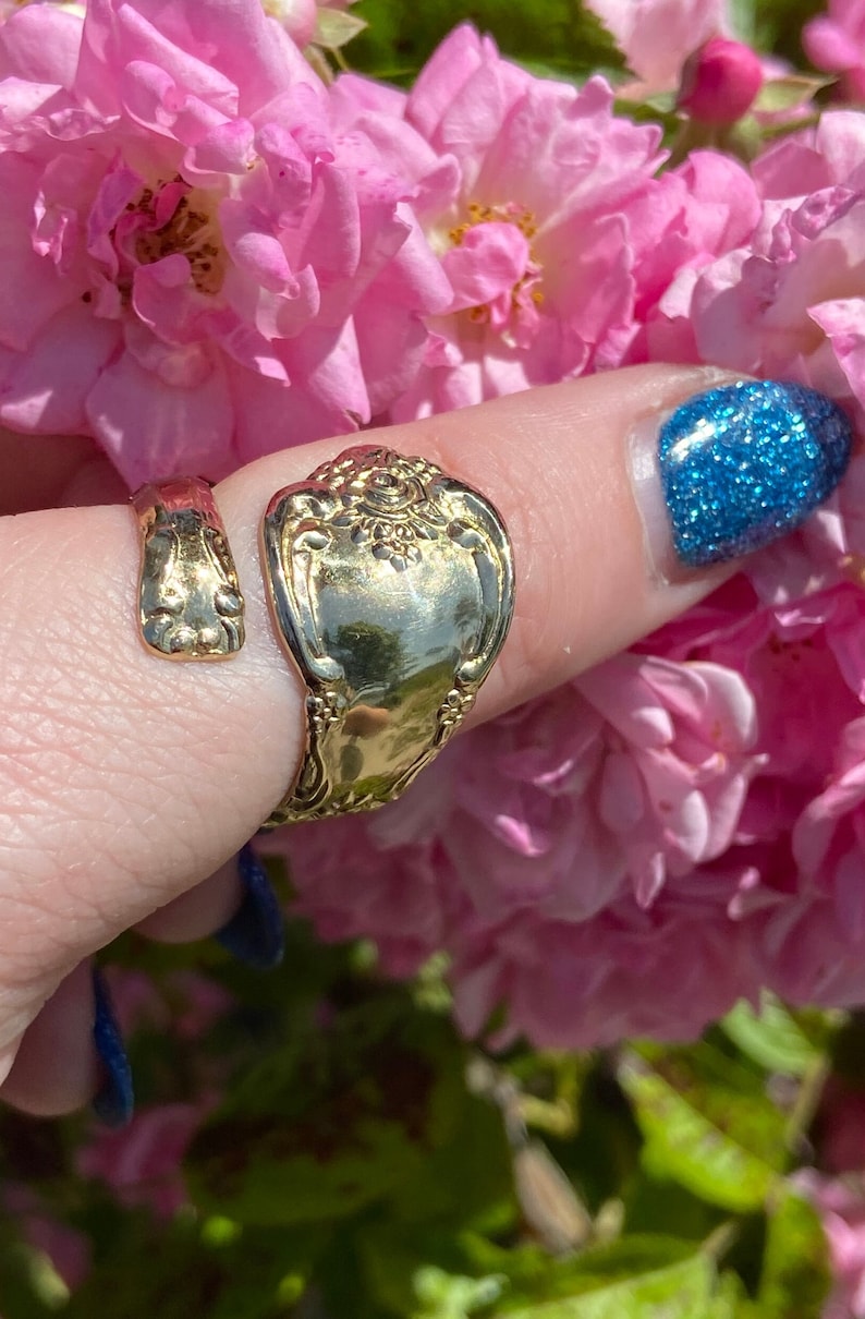 May include: A gold-toned spoon ring with an ornate design. The ring is worn on a finger with blue nail polish.