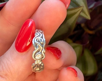 May include: A silver ring with a detailed, intricate design featuring a swirling pattern. The ring is worn on a finger with red nail polish.