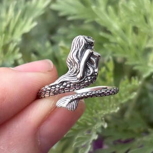 May include: A silver mermaid ring with a detailed design. The mermaid is curled around the ring band with her tail wrapping around the finger.