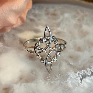 May include: A silver ring with an intricate Celtic knot design. The ring features a central pointed element and swirling patterns, creating a detailed and elegant appearance. The ring is displayed on a light-colored, marbled surface.