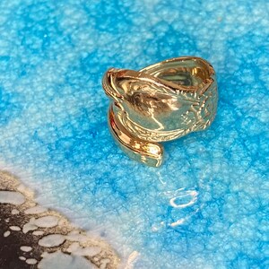 May include: A gold-toned metal ring crafted from a repurposed spoon handle, featuring an embossed floral pattern along the shank. The ring has an open, bypass-style design where the ends of the spoon handle overlap to form the circular band.