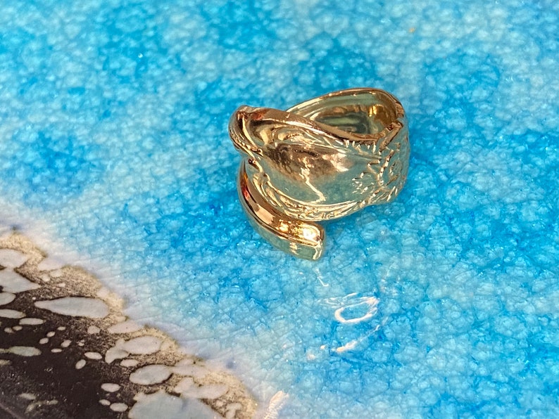 May include: A gold-toned metal ring crafted from a repurposed spoon handle, featuring an embossed floral pattern along the shank. The ring has an open, bypass-style design where the ends of the spoon handle overlap to form the circular band.