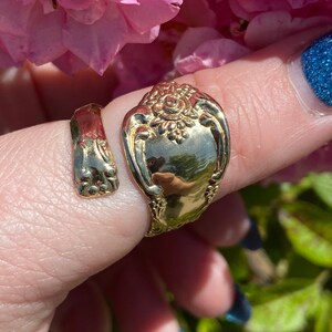 May include: Gold-toned spoon ring with a floral design. The ring is worn on a finger with blue nail polish.