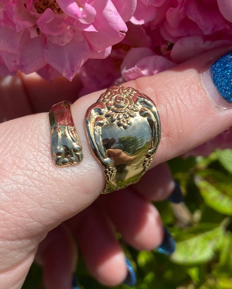 May include: Gold-toned spoon ring with a floral design. The ring is worn on a finger with blue nail polish.