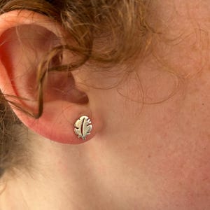 May include: A silver-colored feather stud earring is visible on an ear. The earring is small and detailed, with a cutout design. The ear is next to a person's face, with curly brown hair.
