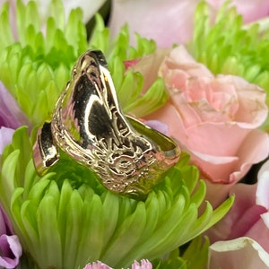 May include: A gold-toned metal ring featuring an open-ended design with a polished surface and an embossed sun motif on the side. The ring is positioned among green chrysanthemum petals and light pink rose petals.
