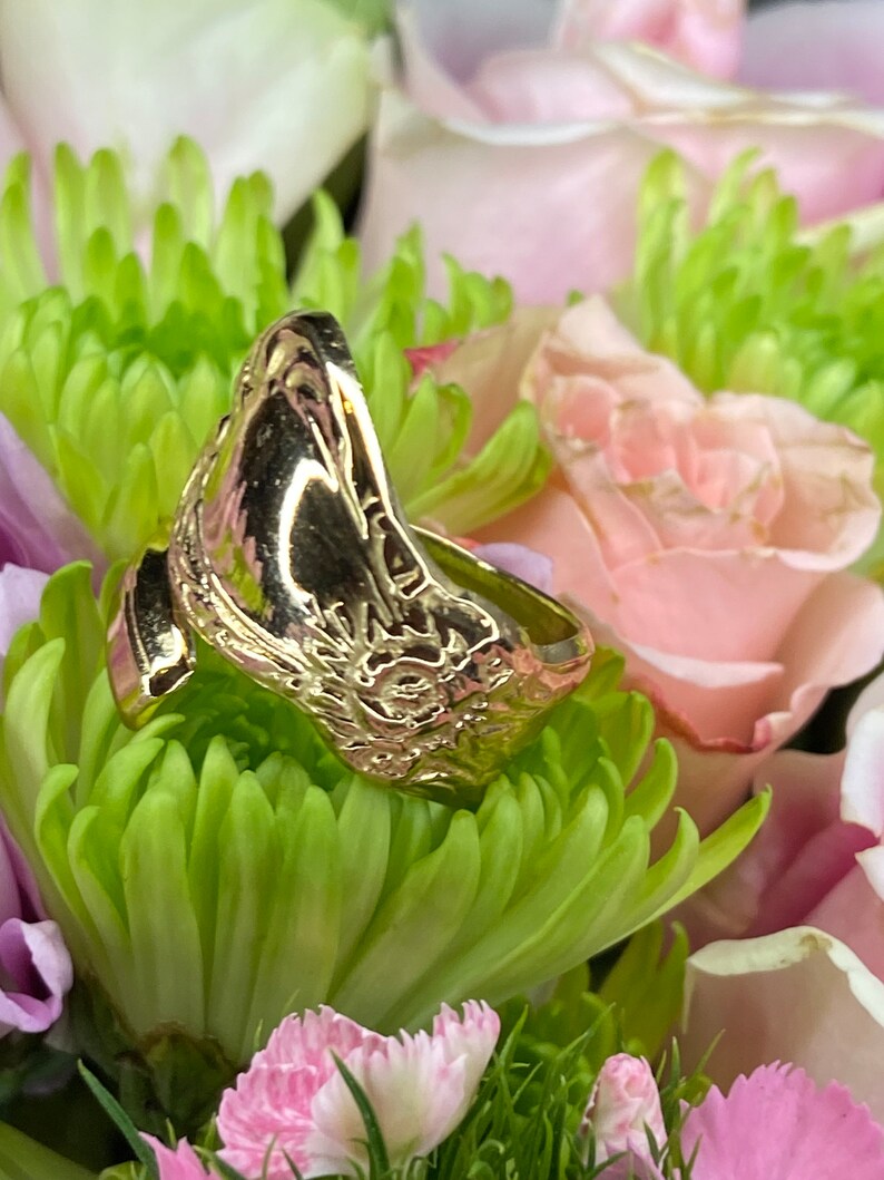 May include: A gold-toned metal ring featuring an open-ended design with a polished surface and an embossed sun motif on the side. The ring is positioned among green chrysanthemum petals and light pink rose petals.