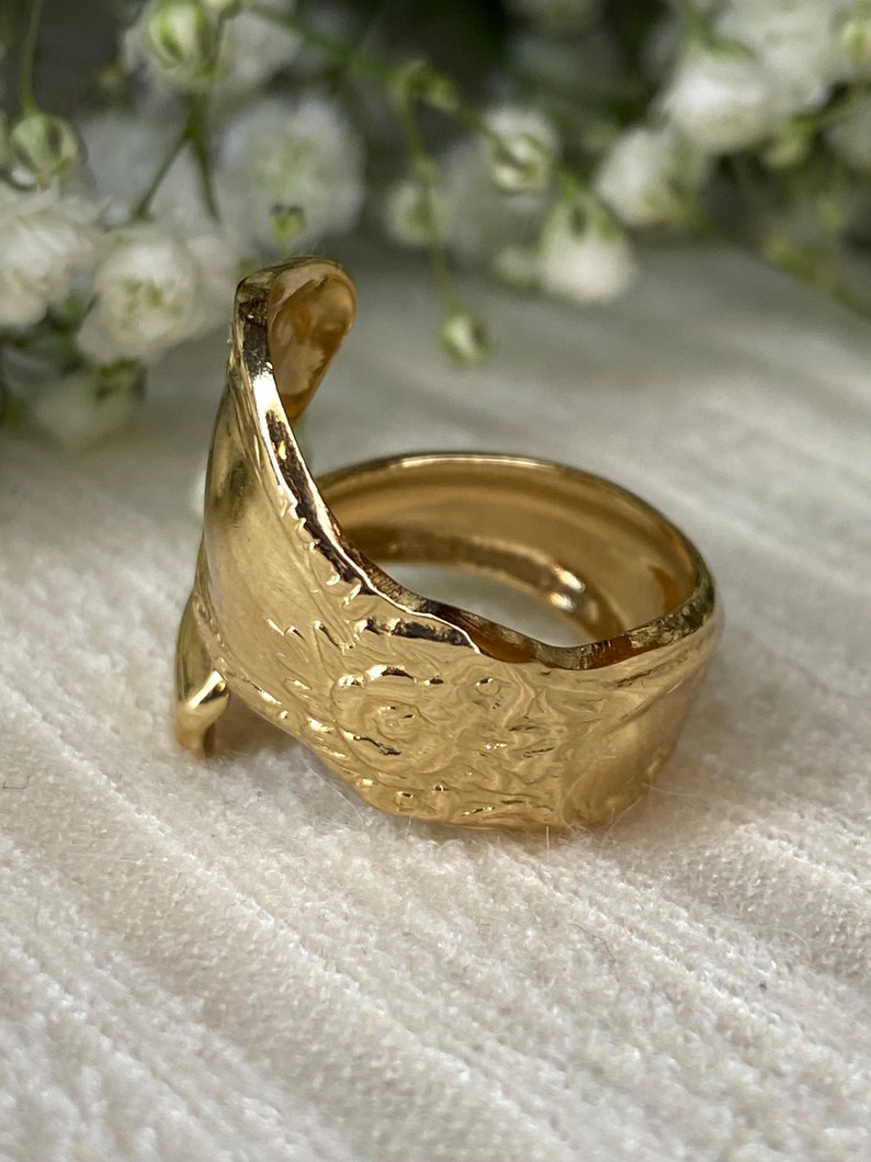 May include: A gold-toned ring crafted from a repurposed spoon, featuring an open-ended design. The band displays a detailed, embossed floral pattern along its exterior surface, with the curved handle of the spoon forming the ring's structure.