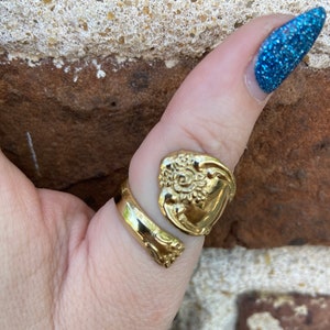 May include: A gold-toned ring made from a repurposed spoon with a floral design. The ring is worn on a person's thumb.