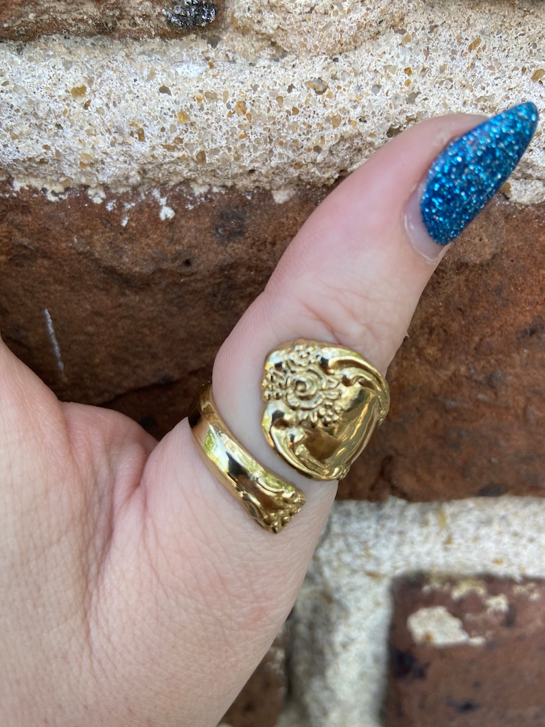 May include: A gold-toned ring made from a repurposed spoon with a floral design. The ring is worn on a person's thumb.