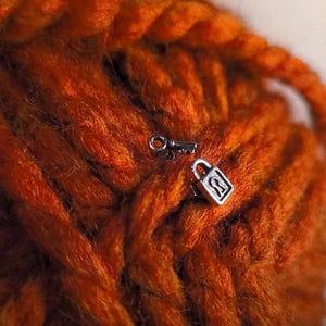 May include: A small silver-toned metal padlock charm featuring a visible keyhole design, positioned alongside a tiny matching silver-toned key charm, both resting against a textured background of thick, vibrant orange yarn fibers.