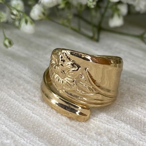 May include: A gold-colored spoon ring with a floral design. The ring is made from a spoon and has a wrapped design. The ring is on a white surface with white flowers in the background.
