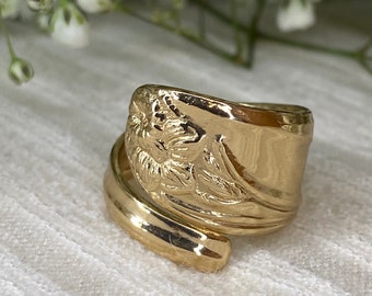 May include: A gold-colored spoon ring with a floral design. The ring is made from a spoon and has a wrapped design. The ring is on a white surface with white flowers in the background.