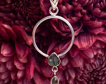 May include: A silver pendant necklace with a circular design and two green gemstone drops. The pendant is hanging against a background of red flowers.