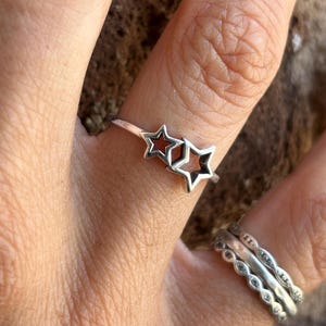 May include: A silver ring featuring two star-shaped cutouts. The ring is worn on a finger, with two other silver rings visible on an adjacent finger. The background is a blurred, neutral tone.