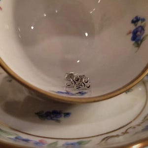 May include: A pair of small, silver-toned metal stud earrings shaped like vintage skeleton keys. The earrings feature a detailed bow, a thin shank, and a bit at the end, resting inside a white porcelain teacup with a gold rim and blue floral pattern.